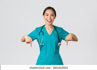 Covid-19, Healthcare Workers, Pandemic Concept. Happy Smiling Beautiful Asian Nurse, Female Doctor In Scrubs Pointing Down, Inviting For Checkup, Showing Hospital Advertisement, White Background