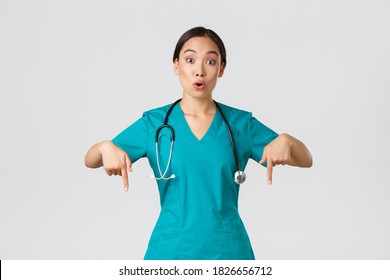 Covid-19, Healthcare Workers, Pandemic Concept. Amazed And Impressed Asian Female Nurse, Doctor In Scrubs Pointing Fingers Down And Looking Questioned, Interested In New Promo, White Background