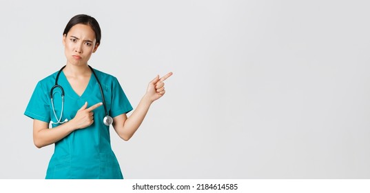 Covid-19, Healthcare Workers, Pandemic Concept. Disappointed And Angry Asian Female Therapist, Nurse In Scrubs Looking Upset At Camera, Sulking And Pointing Upper Right Corner, Wait Explanation