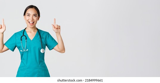 Covid-19, Healthcare Workers, Pandemic Concept. Amused Cheerful Asian Female Nurse, Doctor Or Surgeon In Scrubs Pointing Fingers And Looking Up With Satisfied Face, Standing White Background