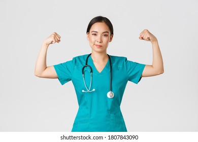 Covid-19, Healthcare Workers And Preventing Virus Concept. Confident Professional Asian Female Doctor, Nurse In Scrubs Smiling Sassy And Flex Biceps, Being Strong, Show-off Muscles