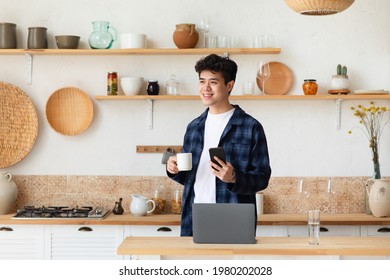 Covid-19 lockdown coffee break and work from home. Smiling asian millennial man holds smartphone, drinks hot tea from cup and looks towards empty space in boho style kitchen interior with laptop - Powered by Shutterstock