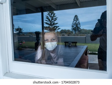 COVID-19 Lockdown. Depressed Lonely Little Girl With Face Mask Looking Through The Window During Quarantine. Sad Sick Child In Self Isolation At Home. Coronavirusu Outbreak And Children Mental Health.