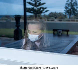 COVID-19 Lockdown. Depressed Lonely Little Girl With Face Mask Looking Through The Window During Quarantine. Sad Sick Child In Self Isolation At Home. Coronavirusu Outbreak And Children Mental Health.