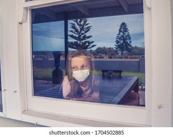 COVID-19 Lockdown. Depressed Lonely Little Girl With Face Mask Looking Through The Window During Quarantine. Sad Sick Child In Self Isolation At Home. Coronavirusu Outbreak And Children Mental Health.