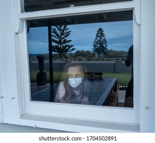 COVID-19 Lockdown. Depressed Lonely Little Girl With Face Mask Looking Through The Window During Quarantine. Sad Sick Child In Self Isolation At Home. Coronavirusu Outbreak And Children Mental Health.