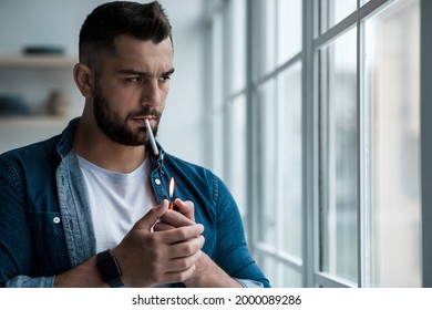 Covid-19 Lockdown, Depression, Stress And Loneliness At Home, Mental Health Problems. Disappointed Sad Millennial Guy Looks Out The Window And Smoke Cigarette In Living Room Interior, Free Space