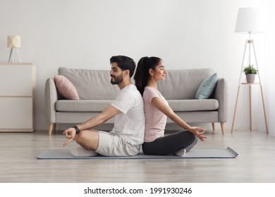 Covid-19 lockdown, workout at home together, active lifestyle. Millennial calm pacified arab man and woman with closed eyes in lotus position, back to back, practice yoga in living room interior - Powered by Shutterstock