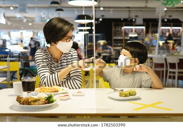 Covid-19 New normal & Physical distancing
concept. An asian mother and son  sit in the cafeteria separated by
a clear acrylic table barrier. Social distancing, Protect against
the coronavirus
disease.