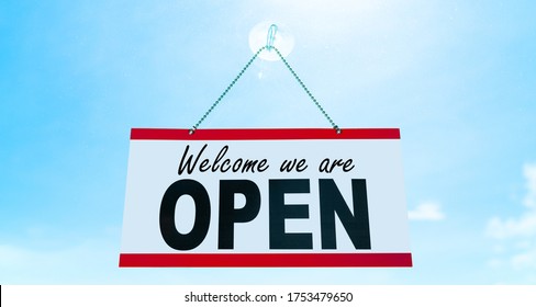 COVID-19 Open Business Sign Saying Welcome We Are OPEN Hanging On Window Storefront Reopening Retail Businesses Opening Again. Sun Blue Sky Summer Background. End Of Confinement.