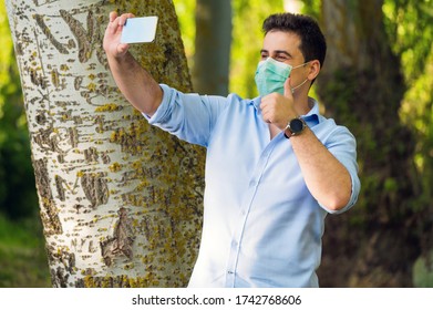 COVID-19 Optimistic Handsome Man Wearing Surgical Mask On Face During Pandemic Coronavirus Disease Showing Thumbs Up In City Park While Doing A Video Call .