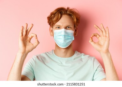 Covid-19 And Pandemic Concept. Handsome Guy With Messy Ginger Hair, Wearing Medical Mask On Face And Showing Okay Signs, Standing Over Pink Background