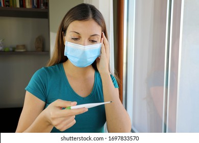 COVID-19 Pandemic Coronavirus Surgical Mask Woman Checking Temperature with Thermometer at Home Symptom of SARS-CoV-2. Girl with mask on face check fever one symptom of Coronavirus Disease 2019.