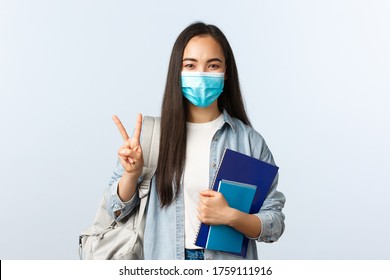 Covid-19 Pandemic, Education During Coronavirus, Back To School Concept. Cute Asian College Student In Medical Mask, Carry Notebooks And Backpack, Show Peace Sign, Starting New Semester