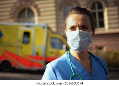 Covid-19 Pandemic. Pensive Modern Medical Doctor Woman In Scrubs With Stethoscope And Medical Mask Outside Near Ambulance.