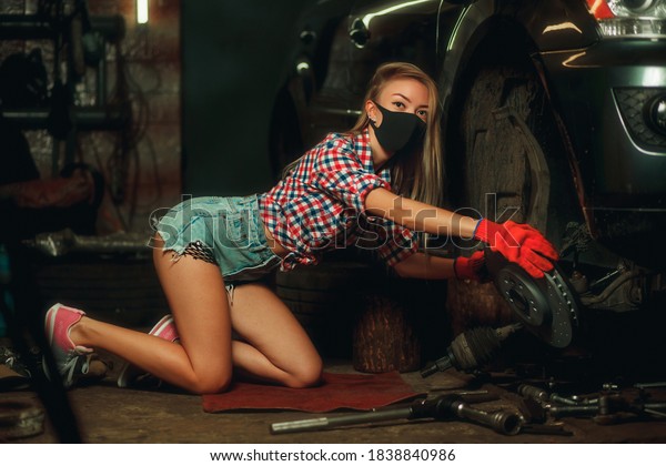 Covid-19 pandemic protection concept. A\
beautiful young girl with a mask on her face is repairing a car at\
a service station. Car wheel rim repair.\
