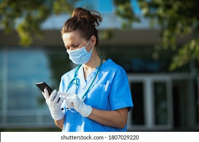 Covid-19 Pandemic. Sad Modern Physician Woman In Scrubs With Stethoscope And Medical Mask Using Smartphone App Outside Near Clinic.