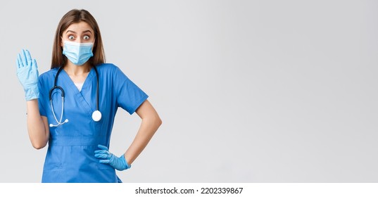 Covid-19, Preventing Virus, Health, Healthcare Workers And Quarantine Concept. Surprised Cute Female Nurse In Blue Scrubs Saying Hello, See Someone In Clinic, Wave Hi, Wear Medical Mask And Gloves