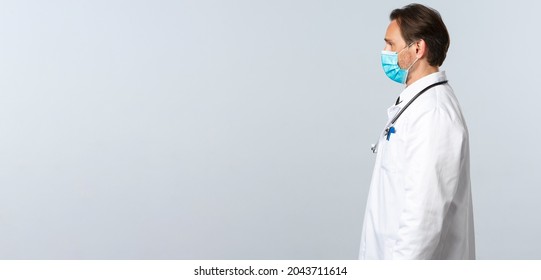 Covid-19, Preventing Virus, Healthcare Workers And Vaccination Concept. Profile Of Professional Doctor At Work Standing In Medical Mask And White Coat, Listening To Patient During Appointment