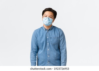 Covid-19, Preventing Virus, And Social Distancing At Workplace Concept. Overworked And Tired Asian Male Employee Looking Reluctant, Wearing Medical Mask, Feeling Bored, Standing White Background