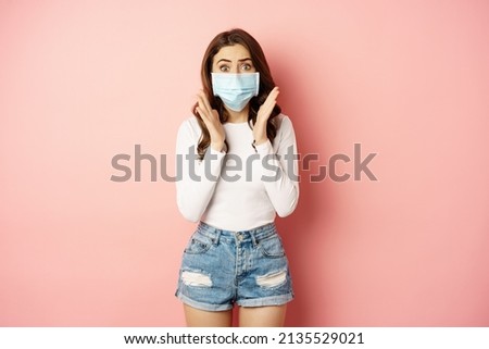 Covid-19 and quarantine concept. Stylish girl in medical face mask, looking worried and shocked at camera, standing against pink background