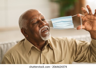 Covid-19 Recovery. Joyful Senior African American Man Taking Off Face Mask Taking A Deep Breath Sitting On Sofa Indoors. Healthcare, End Of Coronavirus Pandemic Concept