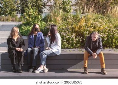 Covid-19, Social Distance Concept. Group Of Talking Women With Protective Face Masks And Man Without Face Mask Sitting In Distance On The Bench During Pandemic Outbreak And Sunny Autumn Day In City