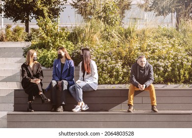 Covid-19, Social Distance Concept. Group Of Talking Women With Protective Face Masks And Man Without Face Mask Sitting In Distance On The Bench During Pandemic Outbreak And Sunny Autumn Day In City