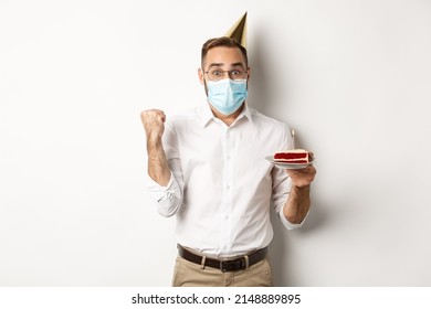 Covid-19, social distancing and celebration. Hopeful happy birthday man in face mask, holding bday cake and rejoicing, standing against white background