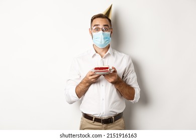 Covid-19, social distancing and celebration. Surprised birthday guy holding bday cake, wearing face mask from coronavirus, white background