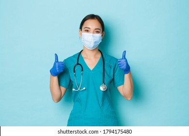 Covid-19, Social Distancing And Coronavirus Pandemic Concept. Optimistic Smiling Asian Female Doctor, Physician In Medical Mask, Rubber Gloves And Scrubs, Showing Thumbs-up, Guarantee Quality Checkup