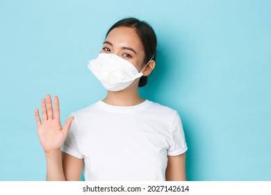 Covid-19, social distancing and coronavirus pandemic concept. Cheerful smiling pretty asian girl in medical respirator saying hello, waving hand in hi, greeting gesture, light blue background