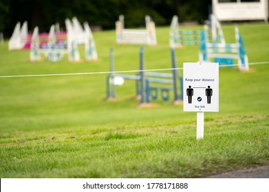 A Covid-19 Social Distancing Sign Staked Into Grass With An Equestrian Show Jumping Outdoor Event In The Background.