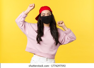 Covid-19, Social-distancing Lifestyle, Prevent Virus Spread Concept. Carefree Excited Asian Girl In Red Cap And Face Mask, Dancing With Hands Raised Up, Having Good Summertime, Yellow Background