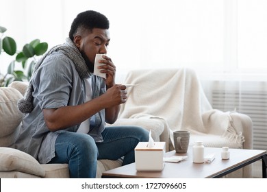 Covid-19 Symptoms. Ill Black Man Coughing And Checking Body Temperature At Home, Feeling Unwell, Sitting On Couch In Living Room