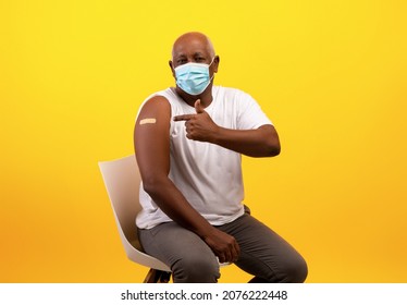 Covid-19 Vaccination. Senior Black Male In Face Mask Pointing At Band Aid After Getting Coronavirus Vaccine Shot On Orange Studio Background. Vaccinated Elderly Man Receiving Anti Covid Injection