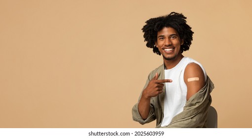 Covid-19 Vaccination. Smiling Millennial African American Guy Pointing At Plaster On His Arm, Young Black Man Got Vaccinated From Coronavirus, Sitting Over Beige Background, Panorama With Copy Space