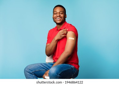 Covid-19 Vaccination. Vaccinated Black Man Showing Arm After Coronavirus Antiviral Injection Sitting With Rolled Up Sleeve On Blue Studio Background, Smiling To Camera. Covid Virus Protection Concept