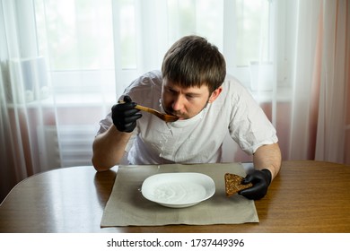 COVID-19. Young Caucasian Man In A White Mask And Black Gloves With Slice Of Bread, Wooden Spoon And A Bowl Of Soup Of Water At A Table In The Kitchen During A Pandemic. Coronavirus In Europe.