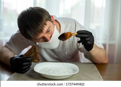 COVID-19. Young Caucasian Man In A White Mask And Black Gloves With Slice Of Bread, Wooden Spoon And A Bowl Of Soup Of Water At A Table In The Kitchen During A Pandemic. Coronavirus In Europe.