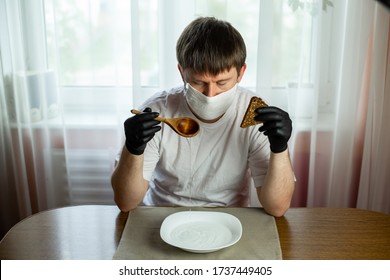 COVID-19. Young Caucasian Man In A White Mask And Black Gloves With Slice Of Bread, Wooden Spoon And A Bowl Of Soup Of Water At A Table In The Kitchen During A Pandemic. Coronavirus In Europe.