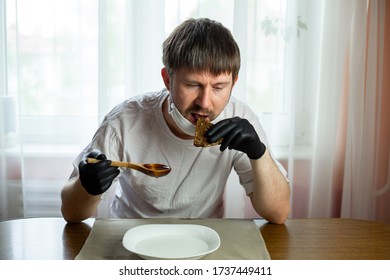 COVID-19. Young Caucasian Man In A White Mask And Black Gloves With Slice Of Bread, Wooden Spoon And A Bowl Of Soup Of Water At A Table In The Kitchen During A Pandemic. Coronavirus In Europe.