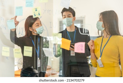 Covid-19. Young People With Face Masks Back At Work In Office After Lockdown.Asian Small Business Startup  Brainstorm Meeting With Laptop And Chart Paper Everyone Mask For Covid19 Protection Corona.