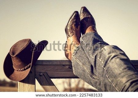 Cowboy boots and hat with feet up on stables gate at ranch resting with legs crossed, country music festival live concert or line dancing concept