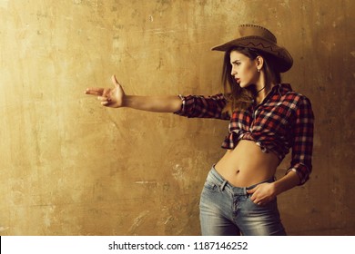 cowboy girl or pretty woman with blond, long hair in stylish hat and red plaid shirt showing finger gun, hand gesture, on beige wall background. Nonverbal communication, gunfighter
