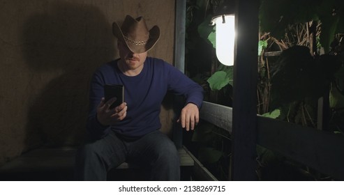 Cowboy Man Communicates Online Using His Phone While Sitting On Porch Of A Village House. Concept Of Lockdowns, Outdoor Living And Farming Activities. Dark Night. Old, Retro Decor, Burlap Background