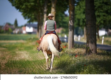 Cowboy rides along the street with a Palomino western horse, photographed from behind.