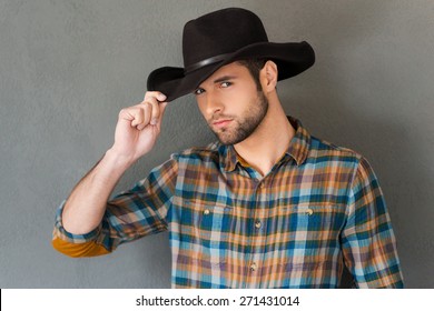 Cowboy style. Handsome young man adjusting his cowboy hat and looking at camera while standing against grey background 