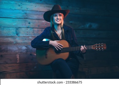 Cowgirl country singer with acoustic guitar. Wearing blue jeans and brown hat. In front of wooden wall.