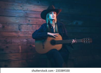 Cowgirl country singer with acoustic guitar. Wearing blue jeans and brown hat. In front of wooden wall.
